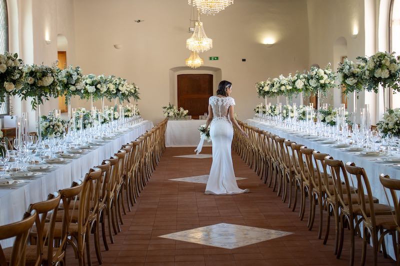 In questa foto una sposa cammina tra due tavoli imperiali decorati con alzata e fiori di colore bianco in un matrimonio firmato da Angela de Patto