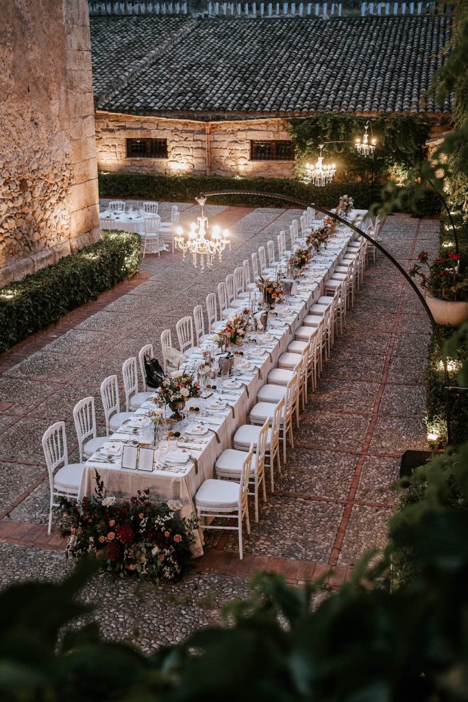 In questa foto inquadrata dall'alto un tavolo imperiale allestito con coppe di fiori di colore bordeaux e rose Playa e rose cappuccino e sormontato da lampadari chandelier