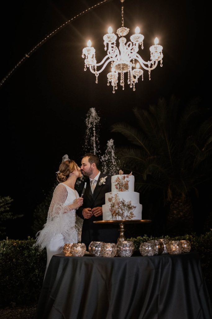 In questa foto due sposi si baciano dietro ad una torta nuziale a tre piani decorata con fiori pressati