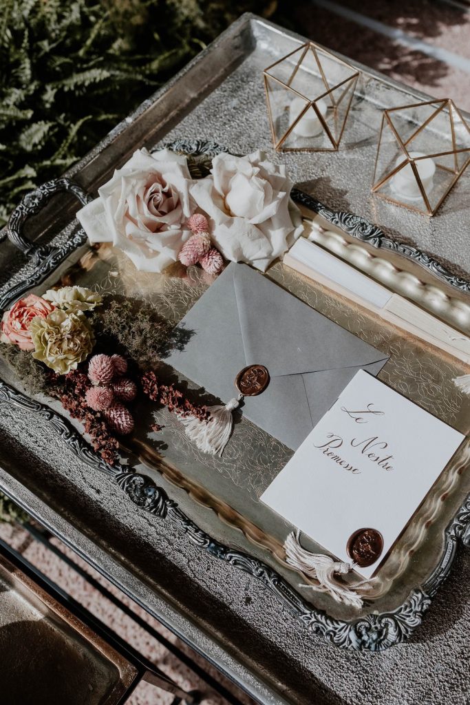 In questa foto posizionati su un vassoio in argento un invito di nozze di colore grigio perla chiuso da un bollino in ceralacca di colore bronzo e un libretto con le promesse di matrimonio en pendant