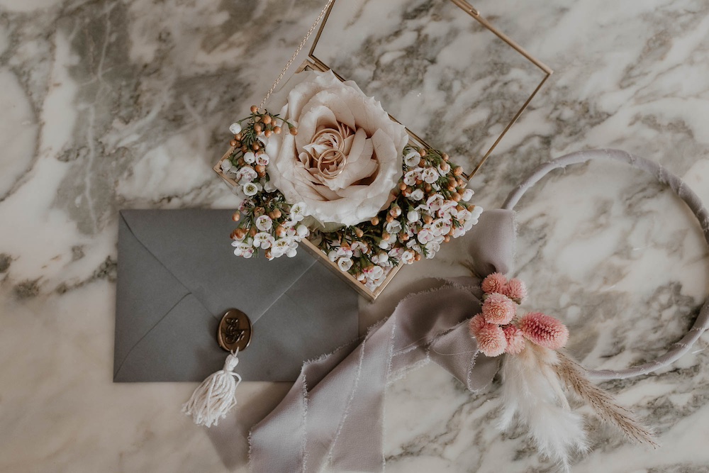 In questa foto una partecipazione di nozze nei toni del grigio antracite, del bronzo e del bianco affiancata al portafedi e all'hoop bouquet nei toni del rosa e del grigio perla