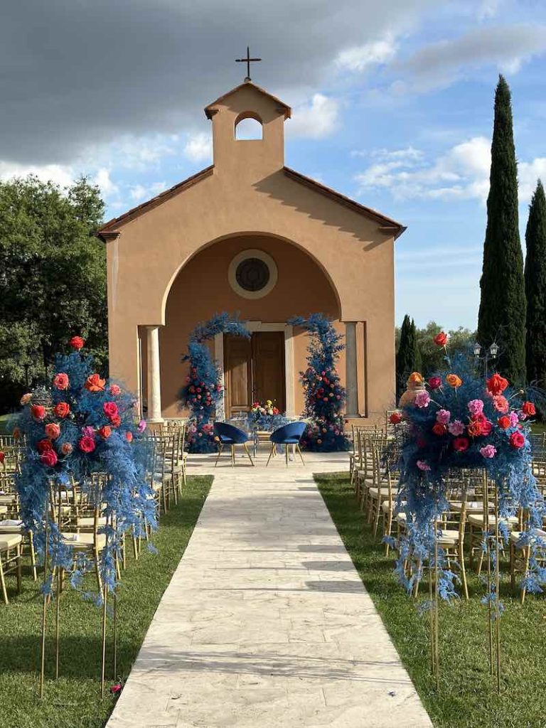 In questa foto un allestimento di una cerimonia di nozze all'aperto realizzato da Barbara Vissani con felce di colore blu elettrico e rose di colore arancione, rosso, giallo e rosa