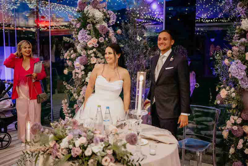 In questa foto due sposi sorridono al tavolo del loro ricevimento di nozze allestito nei toni del lilla mentre la Wedding Planner Barbara Vissani li osserva sorridente