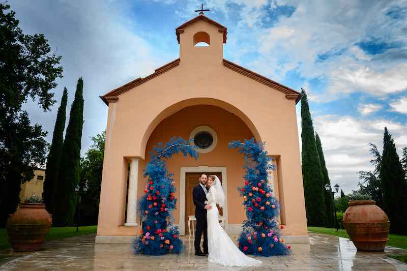 In questa foto due sposi si abbracciano sorridenti davanti ad una  chiesa in un parco la cui facciata è stata decorata dalla Wedding Planner Barbara Vissani con un arco di felce di colore blu elettrico e fiori di colore rosso e arancione