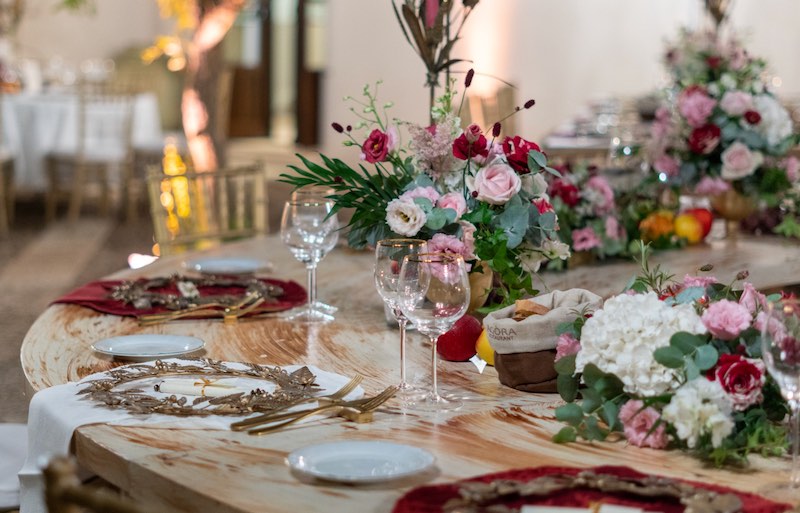 In questa foto un dettaglio della mise en place del Destination Wedding di Vicky e Chris allestito da Rossella Giuffrida al Momentum Resort. La Wedding Planner ha scelto composizioni di fiori di colore rosa, rossi e bianchi con frutta, runner di stoffa di di colore bianco e rosso e sottopiatti a forma di corone d'alloro di colore oro