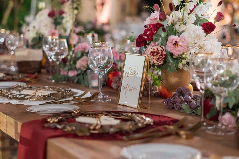 In questa foto un dettaglio della mise en place del Destination Wedding di Vicky e Chris allestito da Rossella Giuffrida al Momentum Resort. La Wedding Planner ha scelto composizioni di fiori di colore rosa, rossi e bianchi con frutta, runner di stoffa di di colore bianco e rosso e sottopiatti a forma di corone d'alloro di colore oro