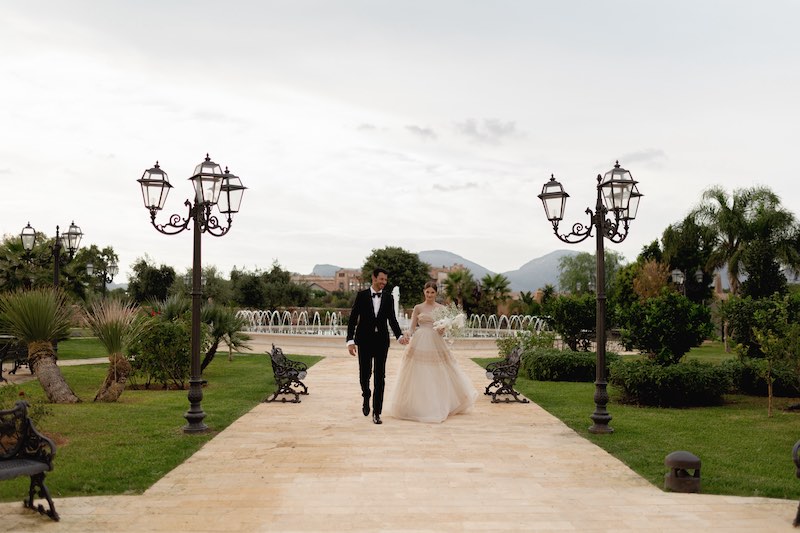 In questa foto due sposi camminano mano nella mano in uno dei viali di Villa del Gattopardo lasciandosi alle spalle la fontana danzante