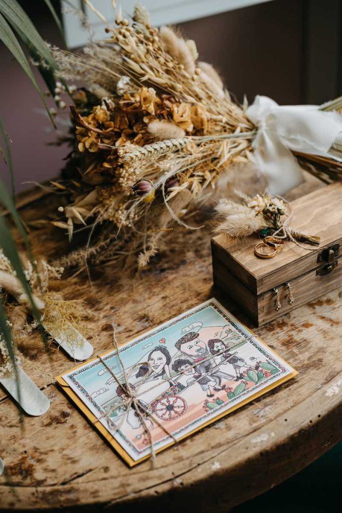 In questa foto la partecipazione di nozze del matrimonio di Tamara ed Enzo con la loro caricatura, un bouquet di fiori secchi e stabilizzati abbinato ad una boutonnière e alle fedi nuziali