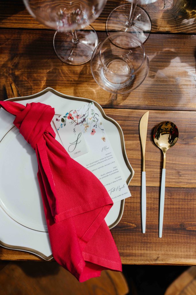 In questa immagine un tovagliolo rosso, annodato sul piatto, e posate dorate. 