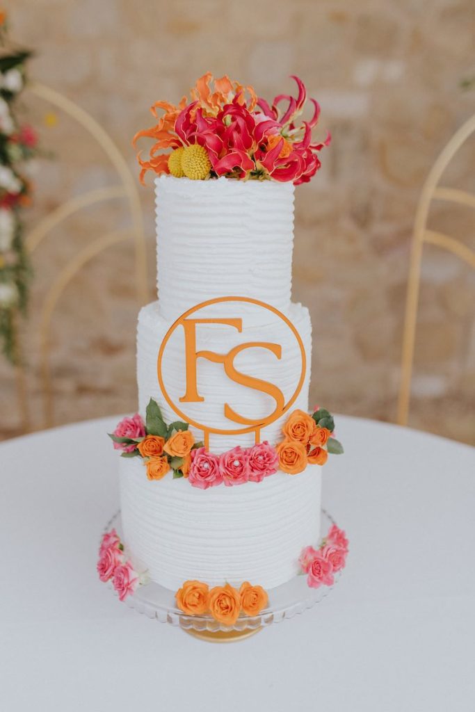 In questa foto la torta nuziale di Francesco e Simona a tre piani decorata con ghiaccia reale di colore bianco, fiori di colore arancione, fucsia e giallo e un topper con le iniziali degli sposi