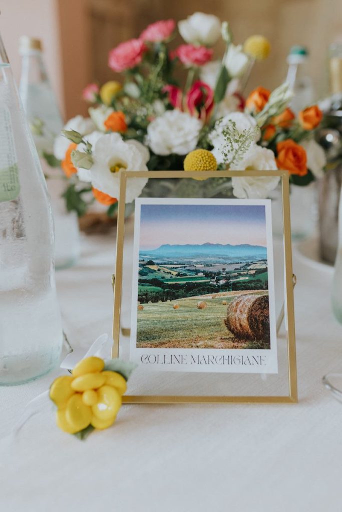 In questa foto uno dei segnatavolo delle nozze di Francesco e Simona composto da cornici dorate nelle quali Elisabetta Alexis ha inserito le immagini dei luoghi più belli delle Marche e dell’Abruzzo e una coppa dorata con fiori di colore arancione, bianco, giallo e fucsia