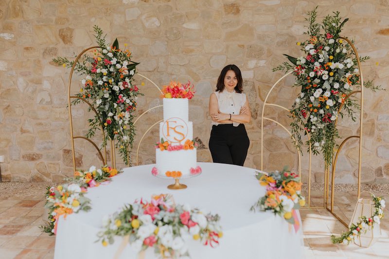In questa foto la Wedding Planner Elisabetta Alexis dietro al tavolo della torta nuziale degli sposi Francesco e Simona. La scenografia è composta da un backdrop di pannelli 3D di colore oro decorati con fiori di colore bianco, arancione. fucsia e giallo