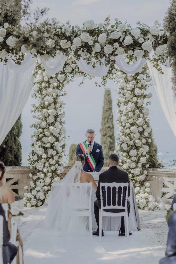 In questa foto due sposi seduti di spalle durante un matrimonio civile allestito sotto ad un gazebo di rose di colore bianco