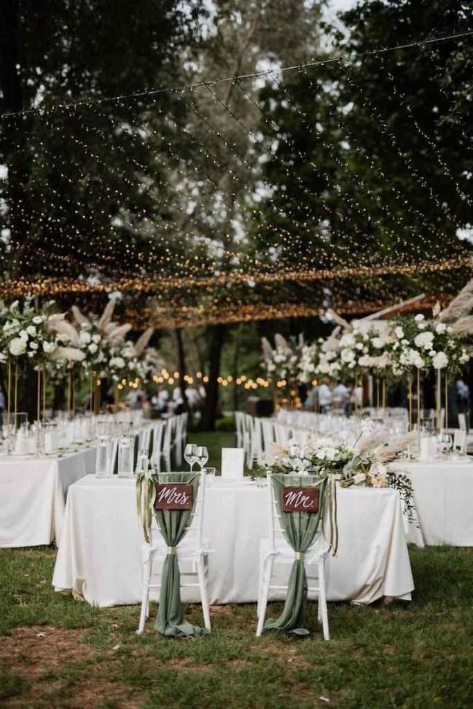 In questa foto un matrimonio allestito su un prato dalla Wedding Planner Nives Malvestiti nei toni del verde e del bianco. In primo piano sono inquadrate le sedie degli sposi decorate con le insegne "Mrs" e "Mr" e con drappi di stoffa di colore verde