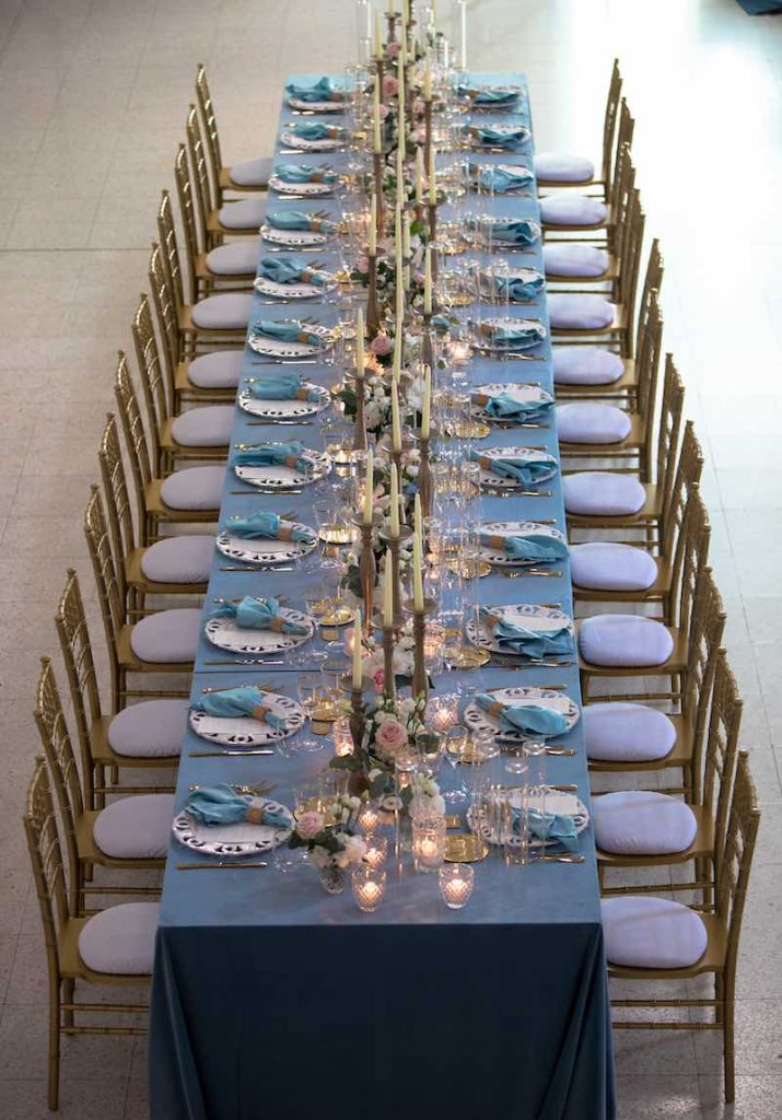In questa foto inquadrata dall'alto un lungo tavolo imperiale allestito con tovagliato di colore azzurro e sedie colore oro, candele e fiori di colore rosa e bianco
