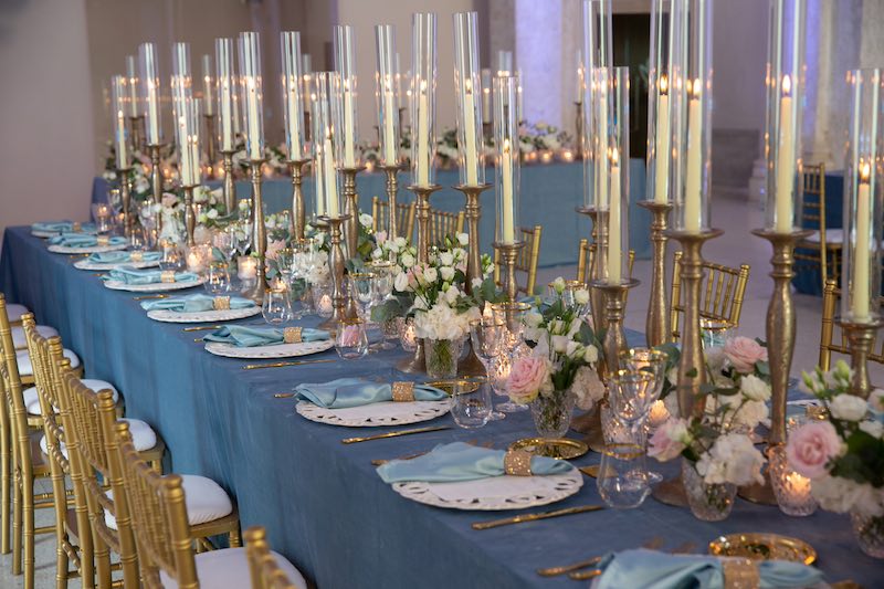 In questa foto il dettaglio di un tavolo imperiale allestito con tovagliato di colore azzurro e sedie colore oro, candele e fiori di colore rosa e bianco