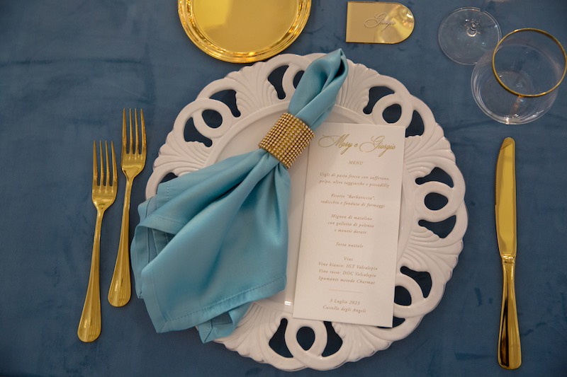 In questa foto il dettaglio di una mise en place realizzata con tovagliato e tovagliolo di colore azzurro, piatto in porcellana e posate di colore oro 