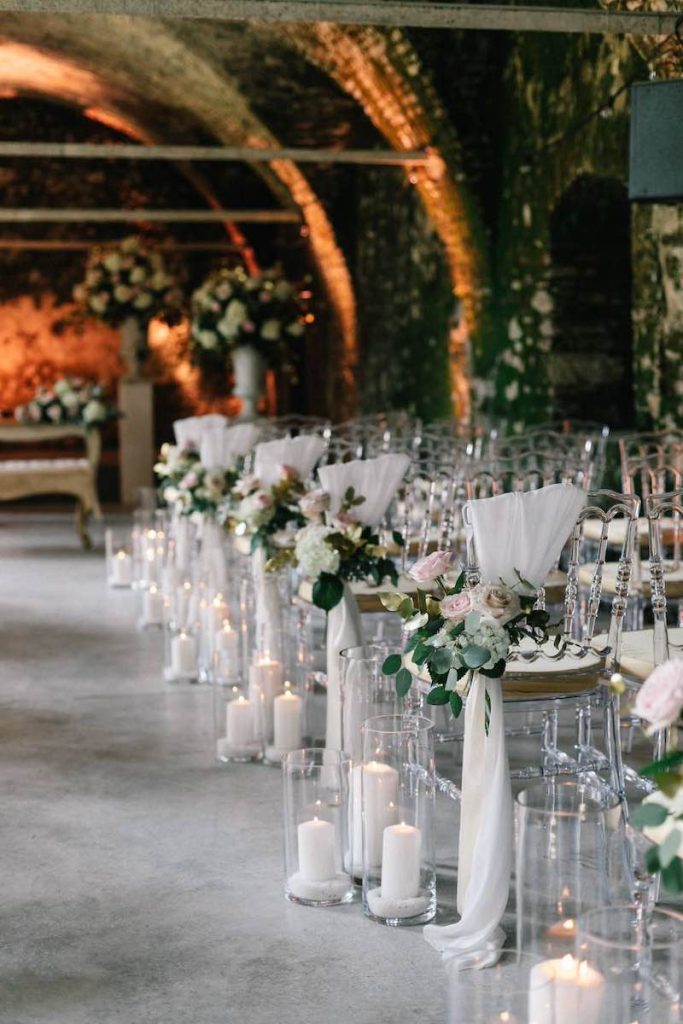 In questa foto il dettaglio dell'allestimento di un matrimonio civile in una sala decorata con rose ed eucalipto legati a un drappo di stoffa di colore bianco a sedie trasparenti e con candele e portacandele di vetro