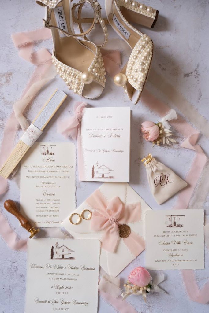 In questa foto scattata dall'alto, la stationery del matrimonio di Roberta e Domenico con partecipazione, menù, libretto messa, sacchetto porta riso, ventaglio, busta, fedi e le scarpe da sposa