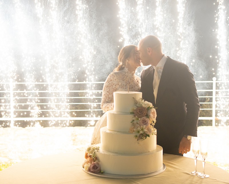 In questa foto due sposi si baciano al taglio della torta nuziale a quattro piani e decorata con fiori. Sullo sfondo sono presenti fuochi d'artificio