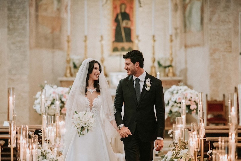 In questa foto di Emiliano Allegrezza due sposi si guardano mentre escono mano nella mano da una chiesa allestita con candele e fiori di colore rosa e bianco
