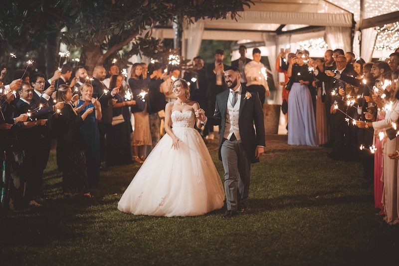 In questa foto di Emiliano Allegrezza, due sposi camminano mano nella mano su un prato mentre ai lati gli invitati tengono in mano le stelline luminose