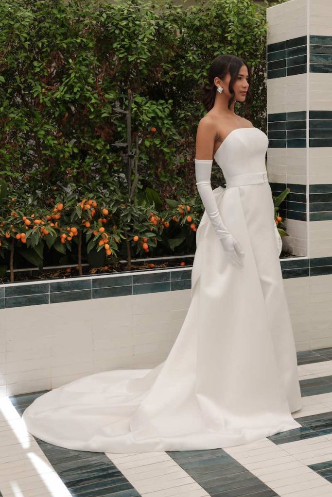 In questa foto una modella indossa un abito da sposa trasformabile con silhouette a sirena e sopragonna. Il look è completato con guanti lunghi