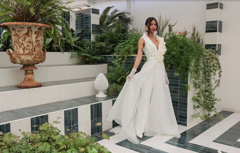 In questa foto una modella indossa un a tuta da sposa con capospalla abbinato e decorato con fiori 3D della collezione Mariù di Atelier Benedetta