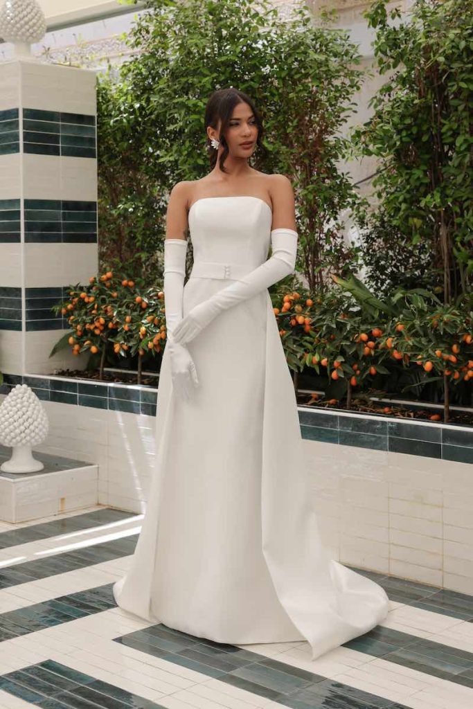 In questa foto una modella indossa un abito da sposa trasformabile della collezione Mariù di Atelier Benedetta con silhouette a sirena e sopragonna. Il look è completato con guanti lunghi