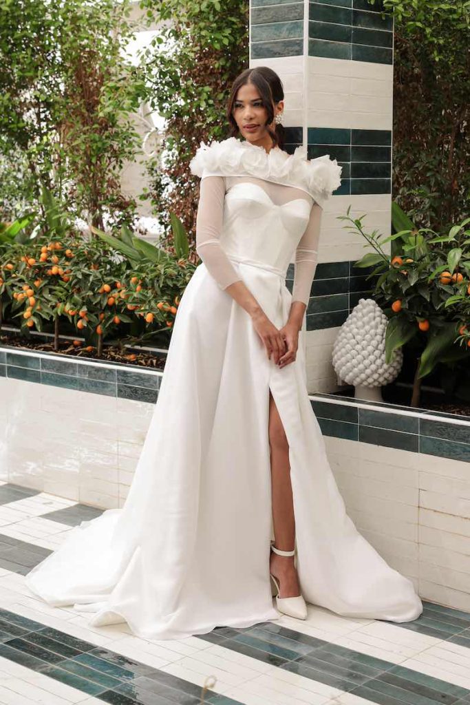 In questa foto una modella indossa un abito ad A con scollo a cuore e coprispalle a maniche lunghe in tulle e fiori 3D della collezione Mariù di Atelier Benedetta