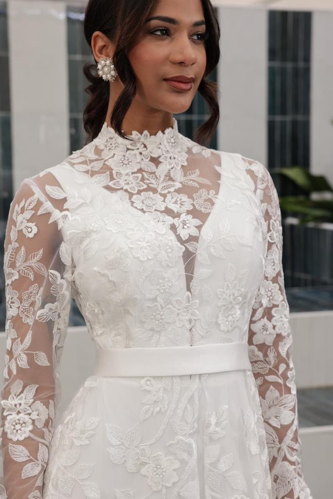 in questa foto una modella indossa un abito da sposa con profondo scollo a V abbinato ad un capospalla in tulle ricamato con pizzo floreale della collezione Mariù di Atelier Benedetta