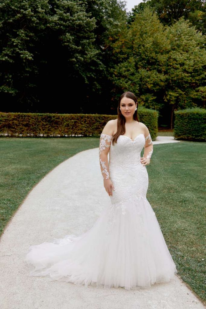 In questa foto una modella  plus size indossa un abito da sposa semi sirena in pizzo e tulle e con maniche a guanto staccabili