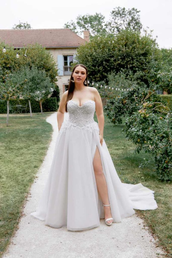 In questa foto una modella plus size indossa un abito da sposa principesco con spacco profondo e corpetto bustier della collezione Sincerity Bridal Primavera Estate 2025