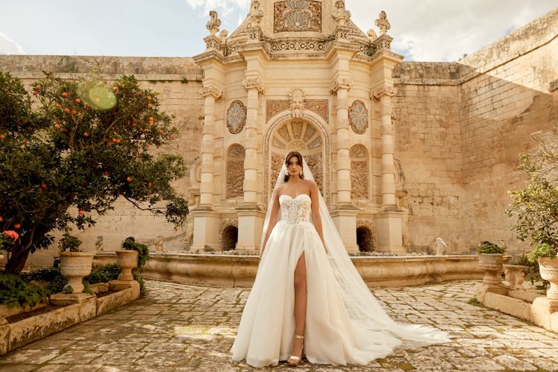 In questa foto una modella indossa un abito da sposa principesco con spacco profondo e corpetto bustier della collezione Sincerity Bridal Primavera Estate 2025