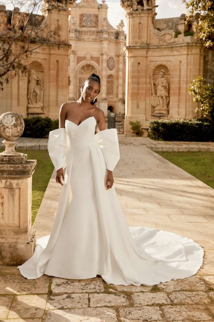 In questa foto una modella indossa un abito da sposa semi sirena in mikado con sopragonna e maniche a fiocco staccabili