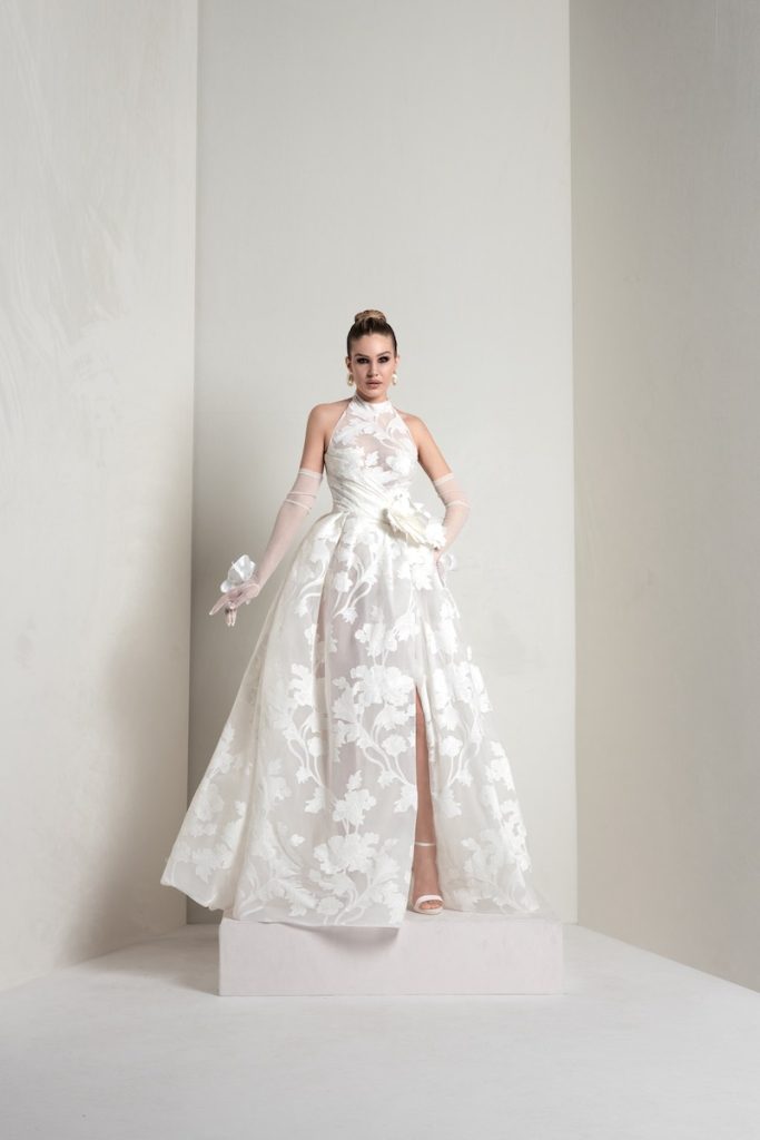 In questa foto, una modella indossa un abito della collezione sposa Markisa 2026 ad A con scollo halter, in organza con maxi fiori ad intarsio e un maxi fiore 3D in vita, con guanti abbinati