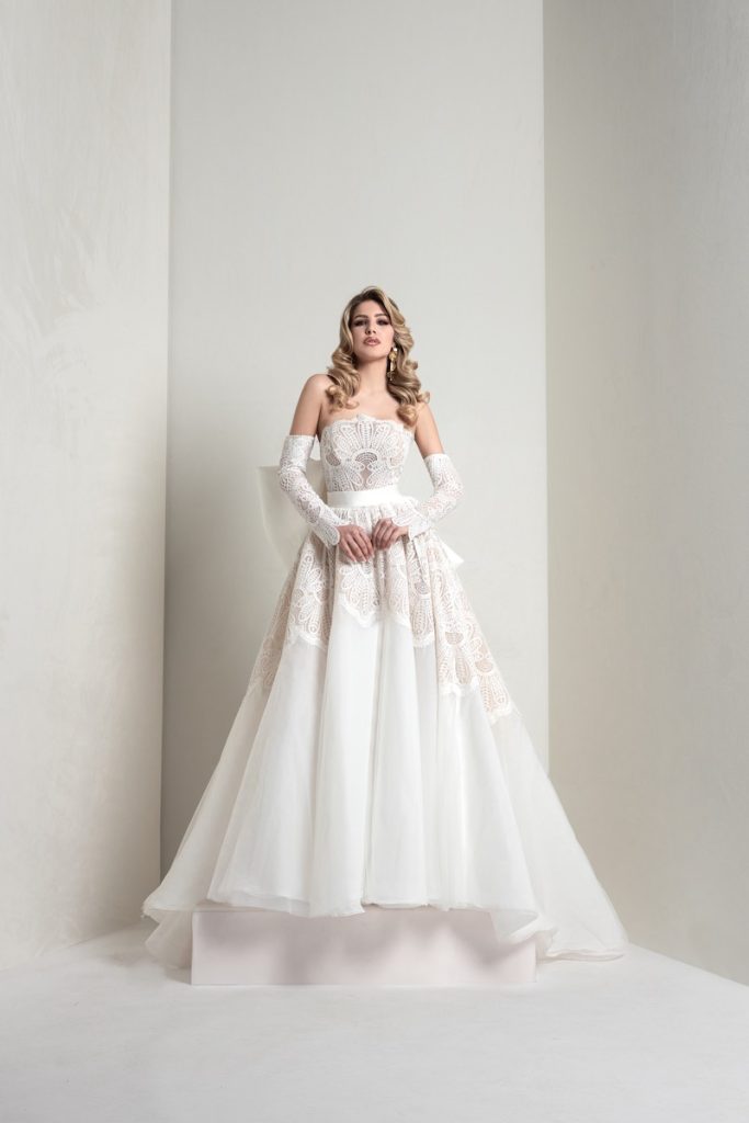 In questa foto, una modella indossa un abito della collezione sposa Markisa 2026 ad A, in pizzo e organza, con manicotti staccabili abbinati e maxi fiocco posteriore