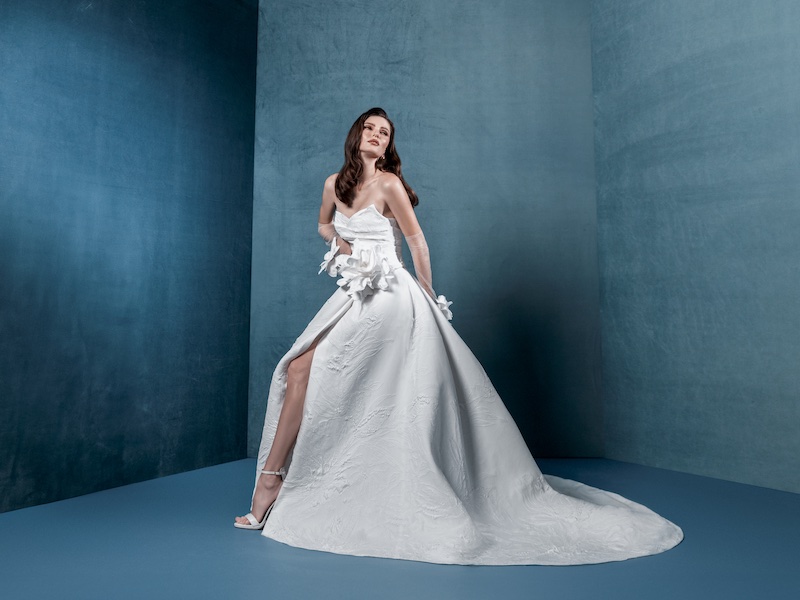 In questa foto, una modella indossa un abito della collezione sposa Markisa 2026 ampio, con spacco frontale e maxi fiore 3D in vita