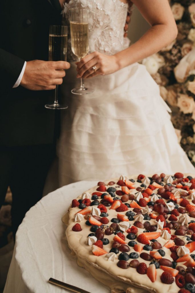 In questa foto due sposi mentre tengono tra le mani i calici di champagne, davanti alla loro torta nuziale ad un piano, bassa, grandissima, circolare e ricoperta di frutta