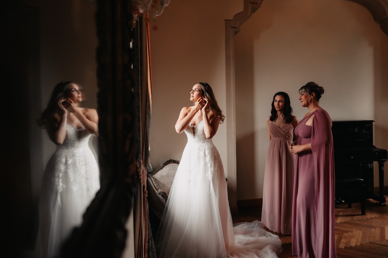 In questa foto di Federico Gritti una sposa mentre si prepara indossando un paio di orecchini mentre due damigelle con abiti color rosa ciclamino la guardano