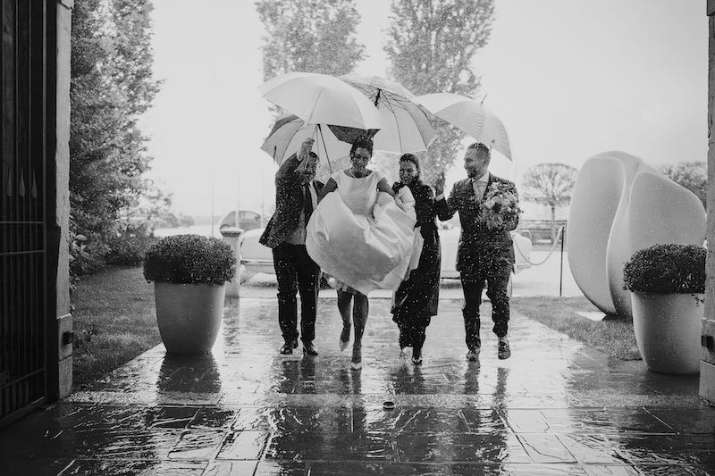 In questa foto  in bianco e nero di Federico Gritti una sposa che sta per entrare in chiesa mentre piove, tiene in mano l'abito e tre persone la coprono con gli ombrelli