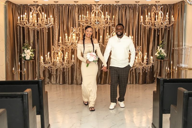 In questa foto, due sposi mano nella mano all'interno della Love Story Wedding Chapel allestista con lampadari di colore oro e un grande sipario di colore bronzo
