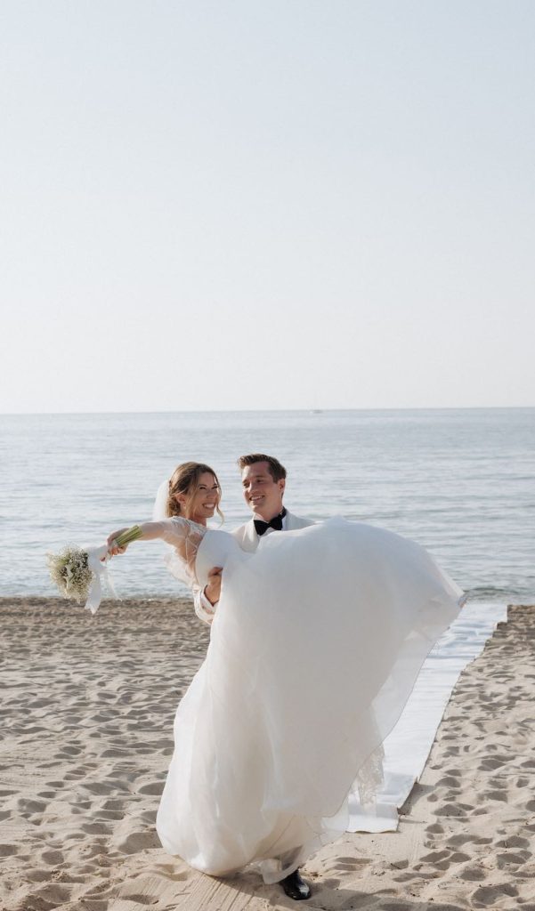 In questa foto, uno sposo tieni in braccio una sposa durante l'ingresso in location sulla spiaggia