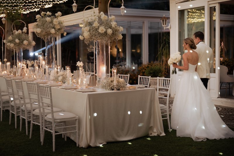 In questa foto, due sposi camminano davanti ad uno dei tavoli del loro matrimonio allestito con fiori, tovagliato e mise en place Total White
