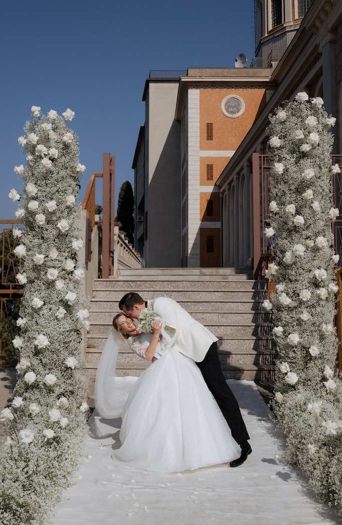 In questa foto, due sposi si abbracciano davanti all'arco di rose bianche e gypsophila con cui è stato decorato l'ingresso della chiesa dal team di Fiorilandia Event Design