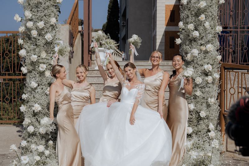 In questa foto, una sposa e le sue damigelle in abiti champagne festeggiano davanti all'arco di rose bianche e gypsophila con cui è stato decorato l'ingresso della chiesa dal team di Fiorilandia Event Design