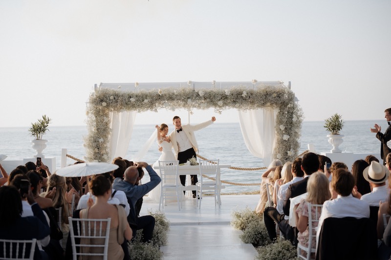 In questa foto, due sposi sotto ad un baldacchino di fiori di colore bianco al termine di un matrimonio simbolico sulla spiaggia firmato Fiorilandia