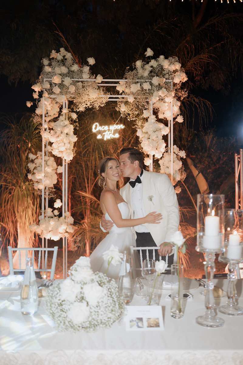 In questa foto, due sposi si baciano davanti alla scritta al neon "Once upon a time" sistemata su un arco di fiori 