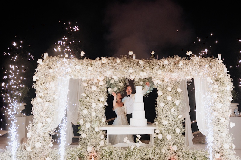 In questa foto, gli sposi al taglio della torta nuziale a piani Total White sotto ad un baldacchino di rose e gypsophila bianche 