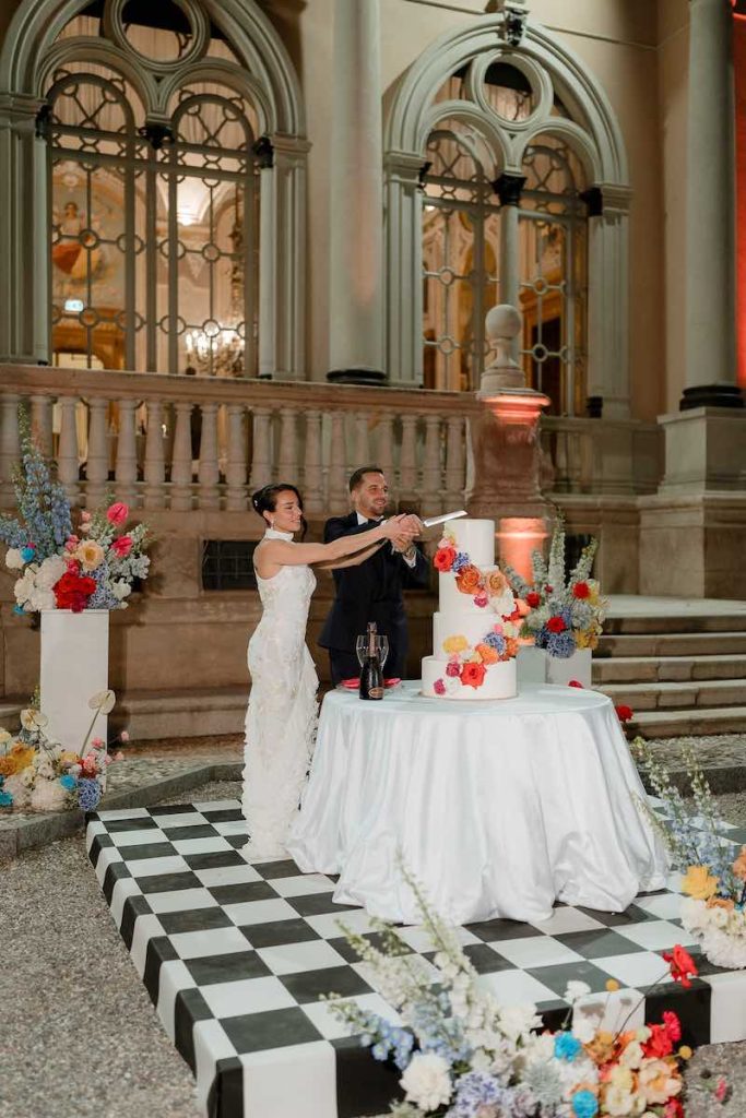 In questa foto, il taglio di  torta a piani per matrimonio di colore bianco, decorata e circondata da fiori colorati